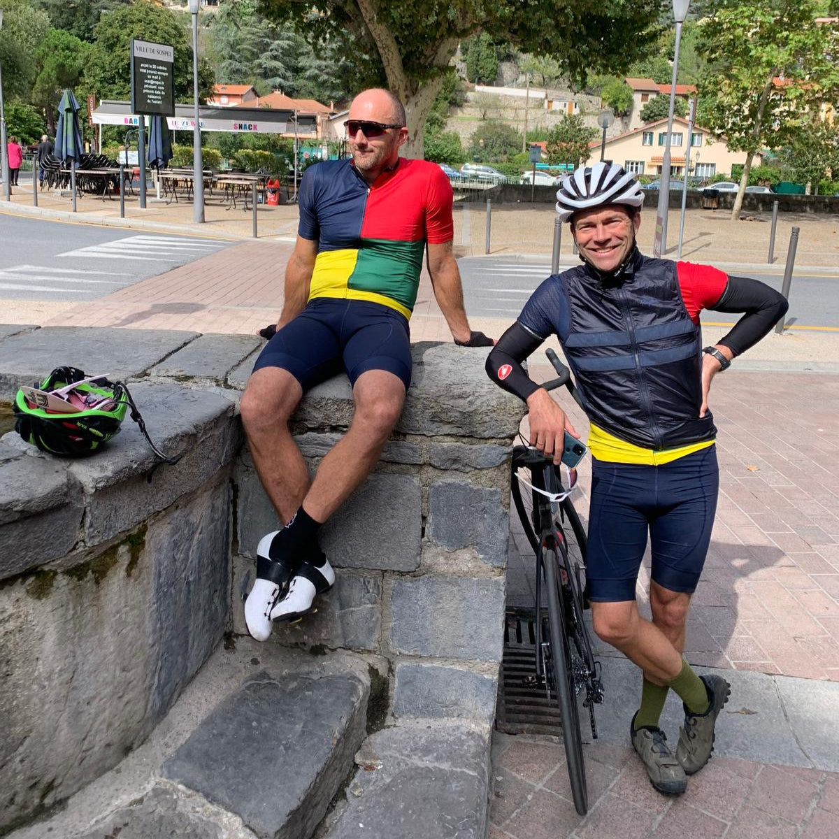 Two cyclists in colorful outfits taking a break on a stone wall with trees and buildings in the background.